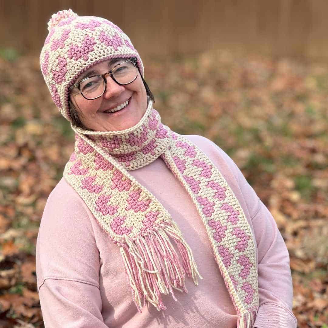 Woman wearing pink and cream knitted hat, scarf, and sweater in autumn setting.